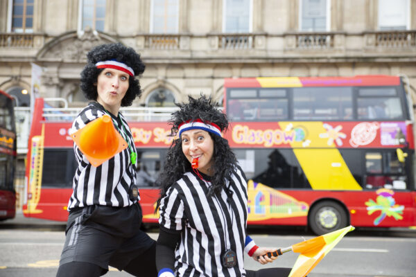 The Refs, two children's street entertainers, are stood in a city square in front of a bus. They are wearing black-and-white-striped referee t-shirts and black curly wigs with striped headbands. One is holding an orange cone towards the camera, the other is holding an orange and yellow flag and blowing a red whistle.