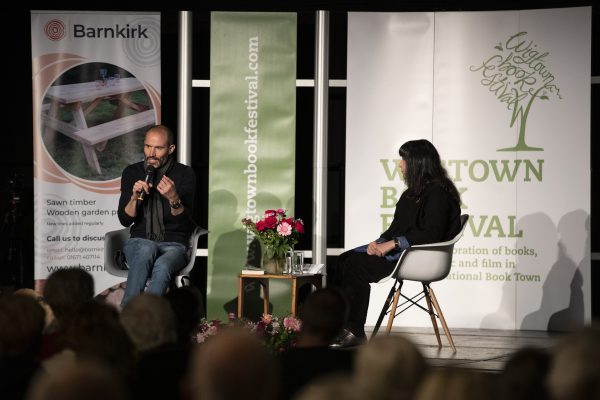 Andrew Cotter and Lee Randall  sitting on stage at a Wigtown Book Festival event. The audience looking on.