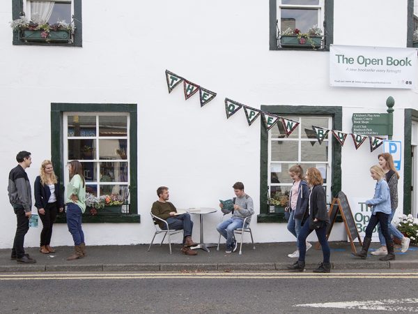 The Open Book bookshop in Wigtown, Scotland's National Book Town. People are walking around outside, two men are seated at the table and chairs reading books. A signpost in green and bunting above it displaying the bookshop's name. Window boxes full of flowers adorn each window.