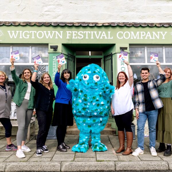 Eight staff members stand beside Big Wig, the big, blue, furry festival mascot, outside the Wigtown Book Festival bookshop. Each each staff holds up a 2023 programme brochure.