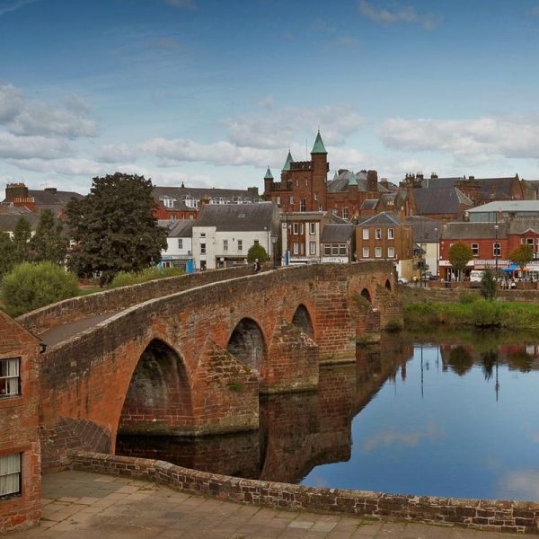 Beside the River Nith at the Old Bridge, Dumfries. A solid low bridge built from red sandstone and with 6 arches.  The town of Dumfries is in the background.