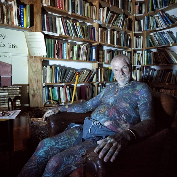 Sandy McNight the tattooed pagan sitting in a comfortable armchair, surrounded by books on bookshelves.