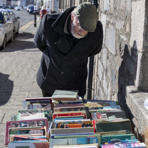 A man is standing outside a bookshop in Wigtown browsing through various books for sale.