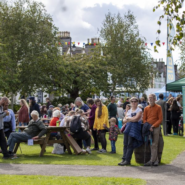 People gathered on a sunny day in Wigtown town gardens during Wigtown Book Festival. A pop up cocktail bar is serving drinks, people are eating at the picnic benches.