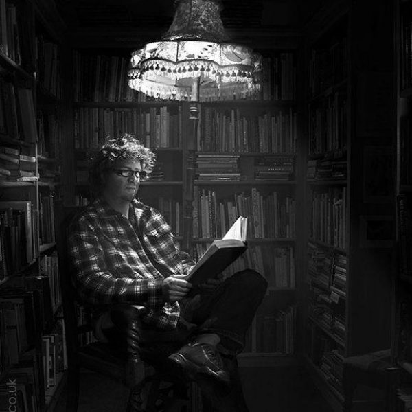 Shaun Bythell sitting in a chair reading a book, an overhead light casts an eerie glow over him. He is surrounded by bookshelves full of books.