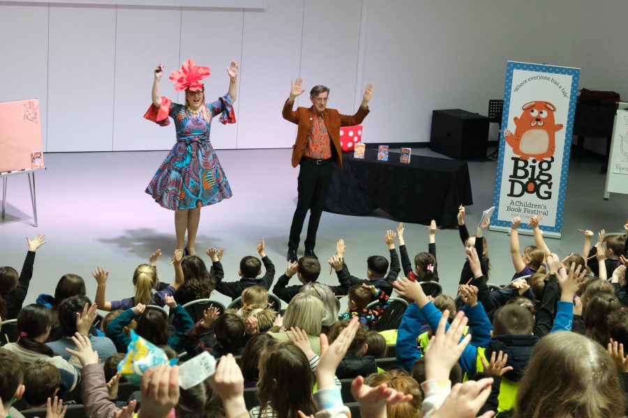 Philip Reeve and Sarah MacIntyre at the Big DoG Schools Gala Day. They are both standing in front of the audience of children with their arms raised up.  Sarah is wearing a bright orange flower hat and flowery dress.