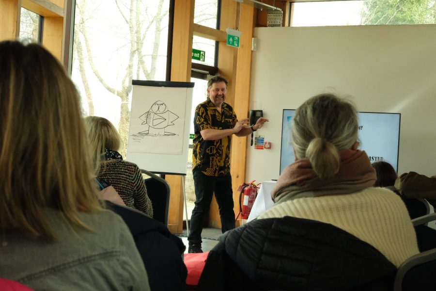 Steven Webb stands in front of a flip chart introducing his book, Peng and Spanners at a Children's Book Festival event. The audience are sitting in front of him.