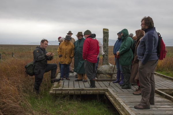 A gathering of people at an outside event in the rain during the Wigtown Book Festival. The event guide is holding various grasses in his hand whilst people look on and listen. They are standing at the Martyrs Stake, Wigtown.