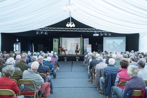 An author event held in the main marquee at the Wigtown Book Festival. Rows of chairs are filled with people listening to the author on the stage. The ceiling is draped with white fabric and a large screen is showing various slide show pictures.