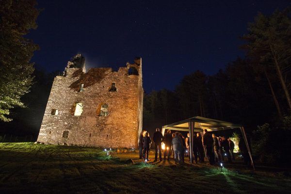 Sorbie Tower lit up in the night sky. Sorbie Tower is a fortified tower house 1 mile east of the village of Sorbie, Dumfries and Galloway, Scotland. A small tent sits to the side with many people underneath talking.