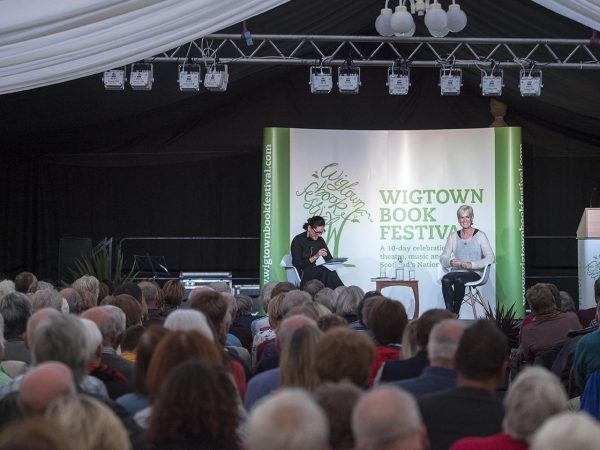 Judy Murray sits on stage with her chairperson at her event at Wigtown Book Festival. The main marquee is full of people sitting listening to her. The ceiling is draped with white cloth, stage lights over their heads.