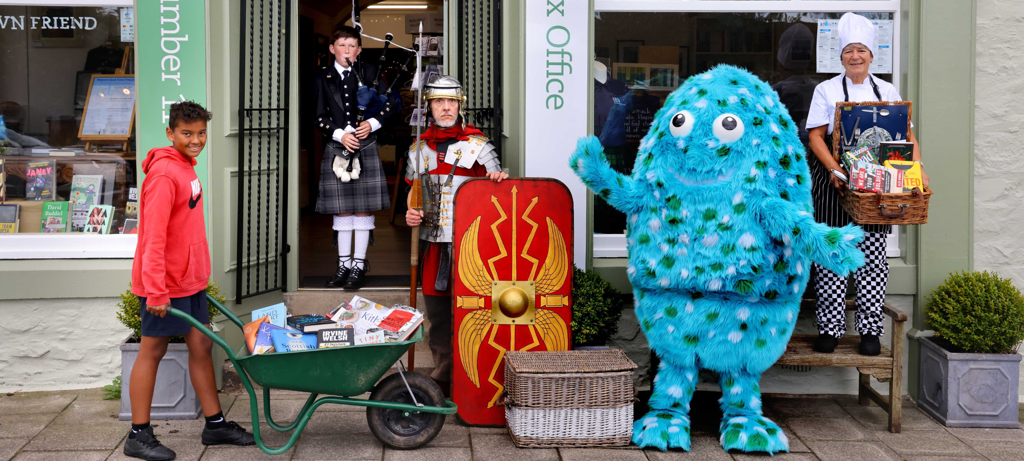 A chef, a roman centurion, a bagpiper and a young person pushing a wheelbarrow filled with books stand outside Number 11 Bookshop