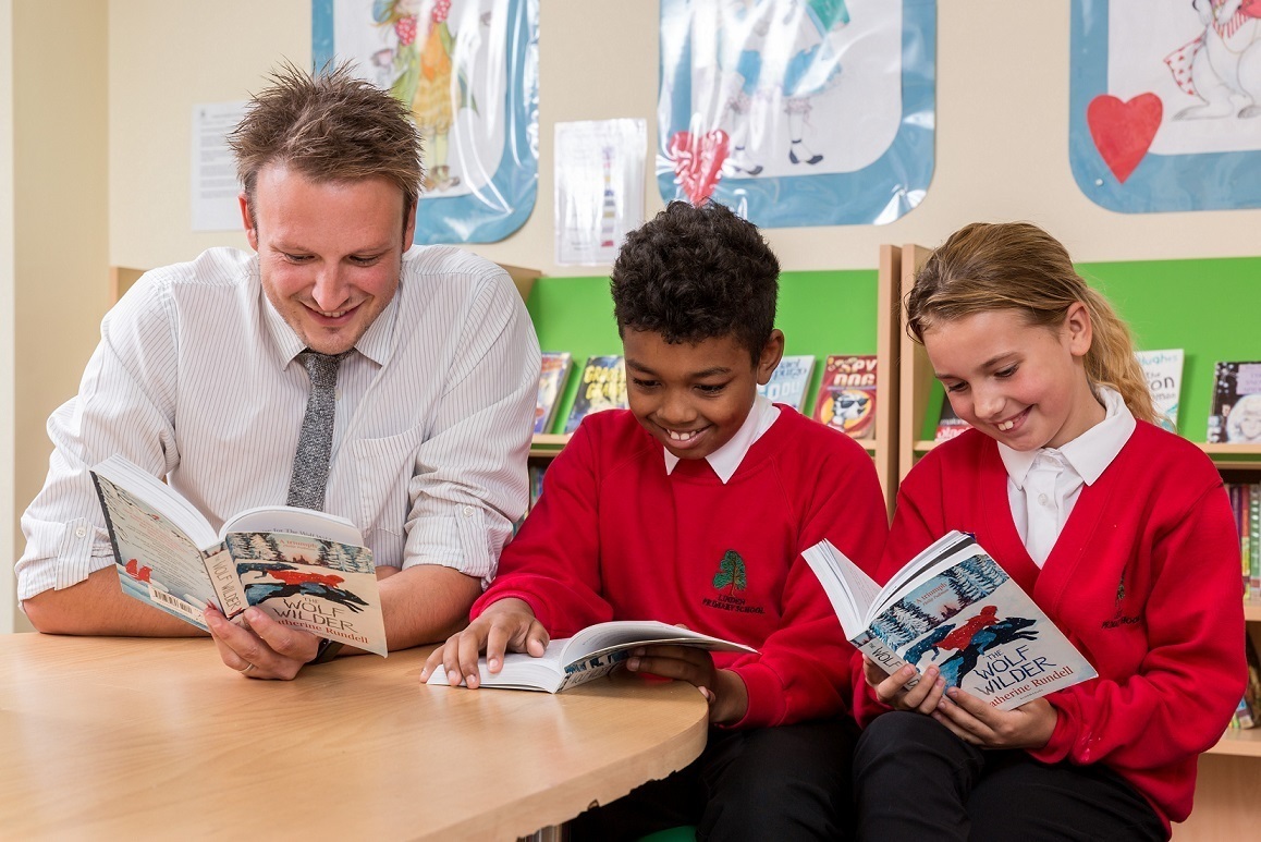 A teacher sitting with two school children in a classroom, each reading a book.